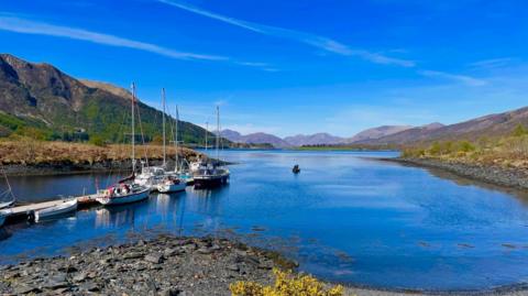 A body of water surrounded by hills with boats moored off the jetty. The sky is clear and the water is bright blue.