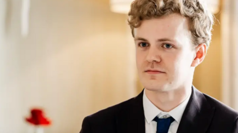 A young man wearing a black suit with a white shirt and a navy tie.