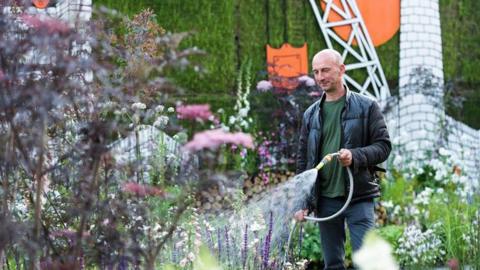 Horticulturalist Nigel Dunnett watering flowers with a hose in a garden. He is wearing blue jeans, a green t-shirt and a black jacket.