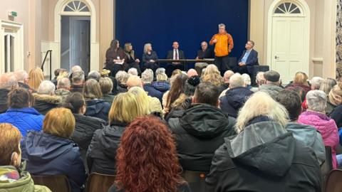 A shot from the back of the hall showing the backs of heads in a crowd. there are people on the stage in the background.