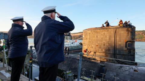 Two men salute as the submarine leaves the naval base. Men standing on the submarine in the distance can be seen to return the salute.