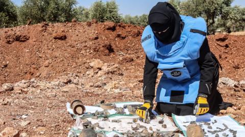 Someone with their face covered and wearing protective clothing sifts through the parts of an unexploded bomb