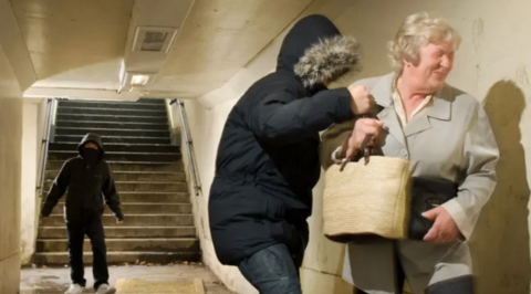 A woman in an underpass is manhandled by a youth who is taking her bag off her. Another youth stands a few feet away. The woman is grimacing. She is wearing a grey jacket and has short grey hair. The youths have their faces covered by furry-edged anoraks.