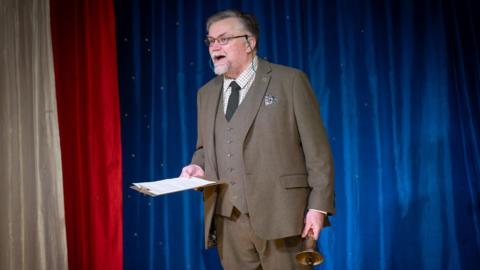 A man with a grey beard, glasses and brown suit standing in front of red, white and blue curtains. He is holding a bell and clipboard. 
