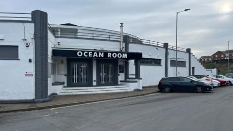 The Ocean Room in Gorleston, with a sign bearing the name above the door. The building is white, with grey art deco pillars. It has three steps to the front entrance, and the end of the building has a staircase built in leading up to what was once a sun terrace.