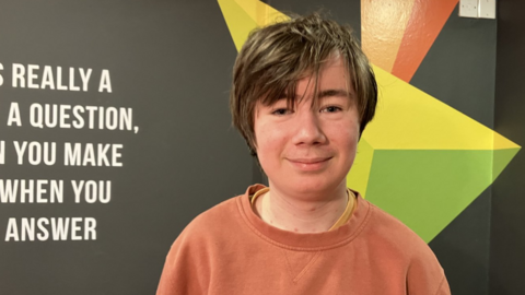 Shows a teenage boy with hair parted from left to right, wearing an orange sweatshirt