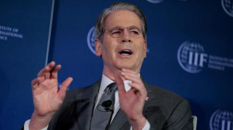 Scott Bessent raises his hands while talking, sitting in front of a dark blue screen.