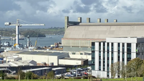 View of an industrial waterfront site with large warehouse buildings, a crane, and docked vessels beside a river or harbour. A modern office building and car park are in the foreground, with cloudy skies overhead.