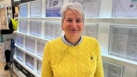 A woman with short white hair wearing a yellow knitted jumper. Behind her are lots of lists of names on white paper which are framed and on a small wall
