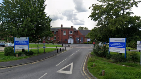 A brick building with a road leading up to it and two blue and white signs either side reading 'Welcome to St George's Hospital and Trust Headquarters' and 'Entrance 1'. Trees can be seen behind the signs and grass all around the road.
