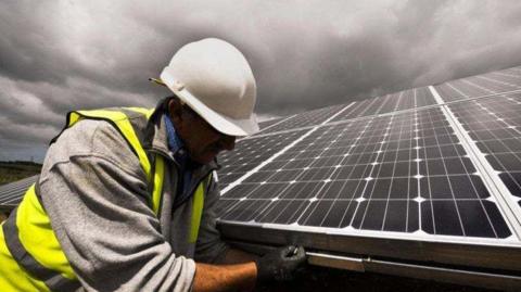 A workman, wearing a white helmet and a yellow hi-vis vest with a grey fleece jumper, blue shirt and black gloves is photographed while fitting a solar panel to a roof on a cloudy day.