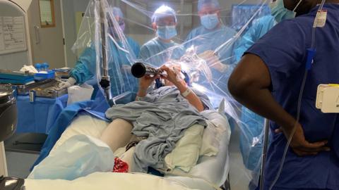 Denise Bacon lying in a hospital bed in a blue gown playing the clarinet