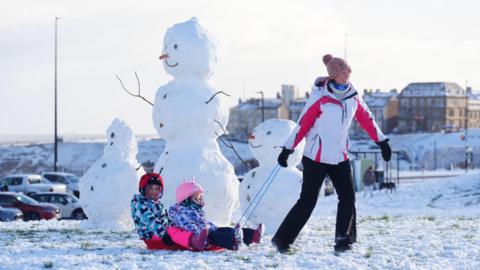 A woman pulls along two children on a sledge with several snowmen in the background.