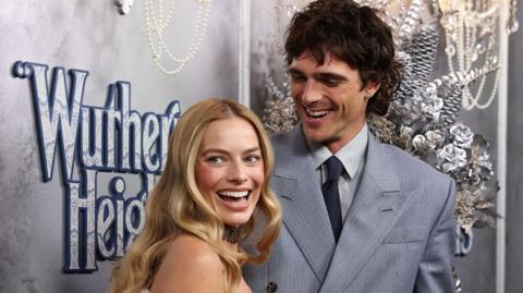 Two smiling celebrities, captured mid-laugh, stood in front of premiere branded background for the Wuthering Heights movie. To the left is blonde haired Margot Robbie looking over her shoulder. To her right is brunette Jacob Elordi looking at the back of her head.