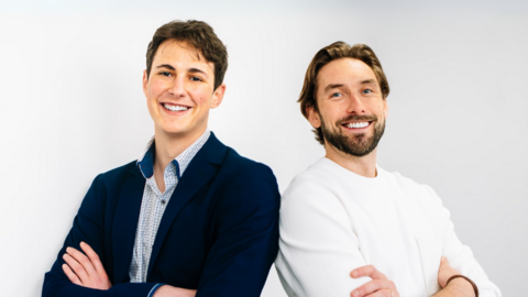 Two men stand smilling shoulder to shoulder, with arms folded. The man on the right has brown hair and is wearing a blue suit and light blue shirt. The man on the right has dirty blonde hair and a beard and is wearing a white sweater.