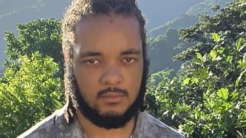 A man with a dark beard and grey shirt looks at the camera. He appears to be stood in a mountainous area with trees and rolling hills in the background.