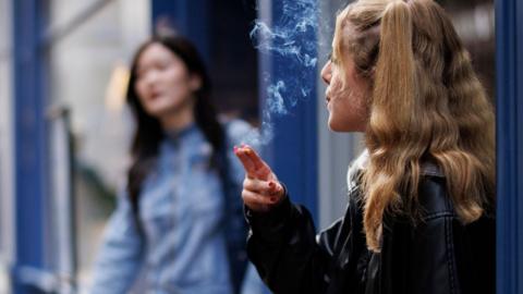 A young woman smokes outside a shop. She has long blonde hair and wears a leather jacket