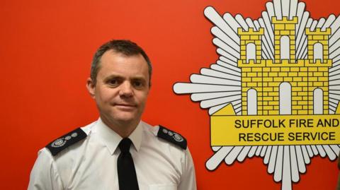 Chief fire officer Jon Lacey standing in front of a red wall with the Suffolk Fire and Rescue Service logo 