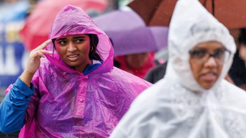 Two people are pictured walking on a street wearing plastic overcoats during a rain shower, on Westminster Bridge in London in June.
