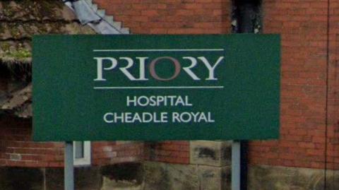 A close up of a green sign set in front of a red brick wall that reads in white writing 'Priory Hospital, Cheadle Royal'