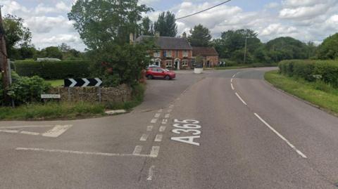 A view looking down the A365 - there is a junction with Spout Lane on the left. There is a bend in the A-road with a traditional red brick pub on the left too. Greenery on either side.