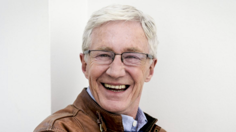 O'Grady smiles at the camera in front of a white wall. He is silver-haired, the picture taken a few years before his death.