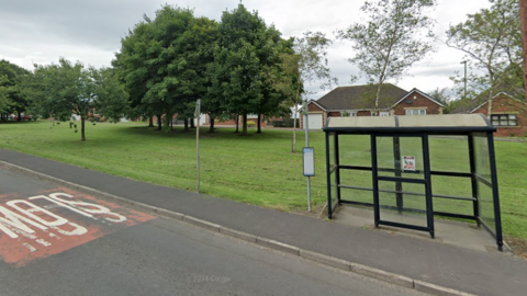 A glass bus shelter in Burnhope. A grass field is between it and brown brick bungalows. 