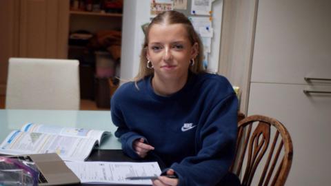 A girl with blonde hair wearing a dark blue sweatshirt with a Nike logo on it. She is sat at a table with school books around her. She is looking at the camera and smiling