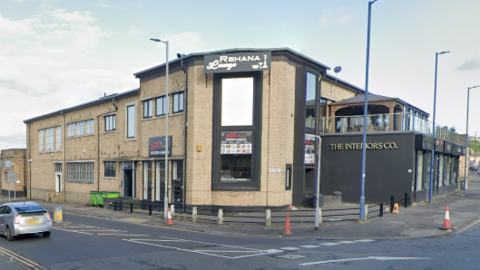 A yellow stone building with a black balcony and large glass windows. A sign reads: "Rehana Lounge". A road passes the venue, with a silver car driving from right to left.