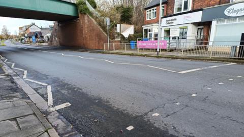 A view of Flamborough Road, Bridlington taken from a footpath. Cracks are visible on the tarmac surface which runs under a green and brick railway bridge. A row of shops is visible on the right of the image and metal safety barriers line the route.