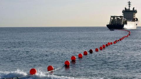 A boat in the distance with a cable connected by orange bollards floating on the sea.