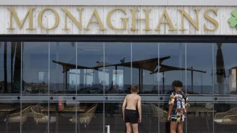 Two boys peering inside Monaghans bar in Costa del Sol. One of them is wearing black shorts and the other is wearing a long shirt covering his shorts.