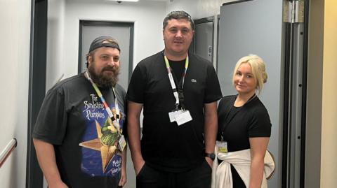 A man in a backwards cap and a Smashing Pumpkins t-shirt stands on the left of a tall man wearing two lanyards and a blonde-haired woman with a jacket tied round her waist on the right. They are standing in a custody suite with cell doors behind them and on the right.