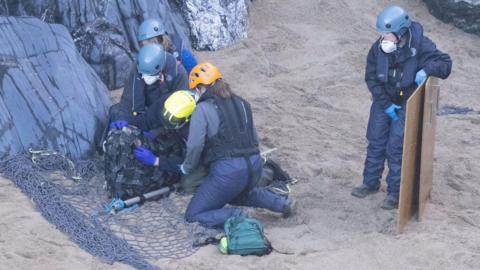 Four people are kneeling around Nanette who is restrained by a cargo net on the beach. A man is standing nearby with a stretcher. 