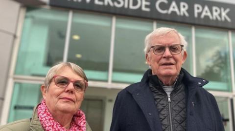 Andrew is wearing a dark blue coat and his wide is wearing a muted green and pink floral scarf