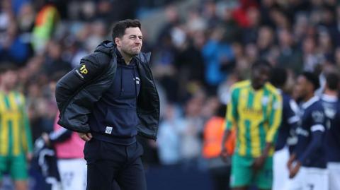 West Brom manager Ryan Mason adjusts the back of his sweatshirt and stares thoughtfully into middle distance after defeat
