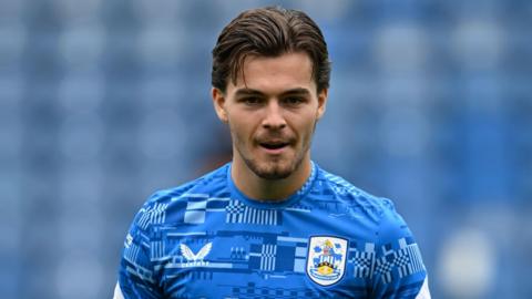 Leo Castledine smiles with mouth slightly open with his gaze slightly down to the ground. He wears a blue patterned training t shirt with the Huddersfield Town badge on it