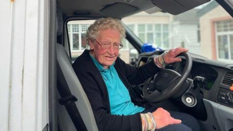 Rosemary Stevenson sits in the driver's seat of a lorry looking out to the side. She is wearing a blue polo shirt, a black cardigan and black glasses. Her hand is on the steering wheel.