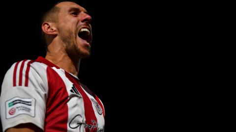 A photo of footballer Matt Lowe celebrating a goal while playing FA Cup football for Brackley Town at Notts County in the FA Cup. Lowe has his mouth open and is looking to his left against a night sky while wearing a red and white striped shirt with a black Adidas logo and the club shirt visible on the chest