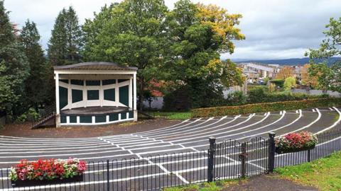 The bandstand, a semi-circular stage with a roof in black and white, sits at the centre of an amphitheatre surrounded by tiered seating. Trees stand behind it, and flowers can be seen hanging on black railings in the foreground.