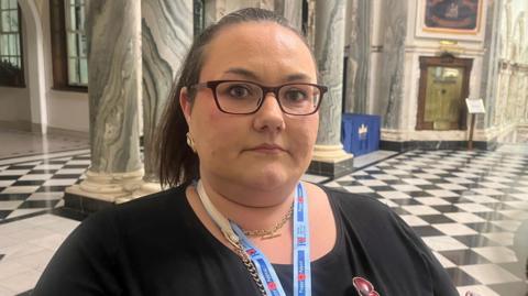 Sarah is standing in large room and in the background there is a black and white tiled floor. Sarah is wearing glasses and has her hair tied back