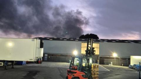 An industrial site with a large plume of black smoke in the distance. The back of a white lorry is to the left of the image.
