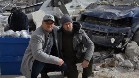 Two men in grey and dark clothing post for a photograph in front of a vehicle which has been hit by a missile 