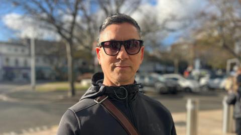 Liam Paterson is standing outside the Government of Jersey building and there are parked cars behind him. He is smiling and has short dark hair. He is wearing sunglasses and a black coat and has a brown leather strap of a bag across his shoulder.