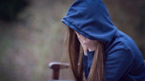 A picture of a young girl with their head in their hands, wearing a blue hoodie. The hood is up and is obscuring their face. 