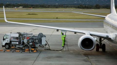 A man in a fluro yellow suit refuels an airplane sitting on an airport tarmac.