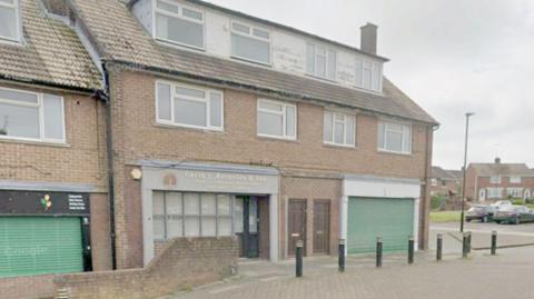 A two storey brown brick building with white dormer windows in the roof. On the ground floor, one half of the building has an office and the other a large green shutter across it. 

