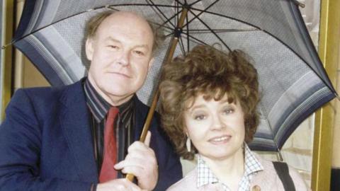An archival photograph of a young Timothy West and Prunella Scales standing close together under a large, open umbrella on a rainy day.