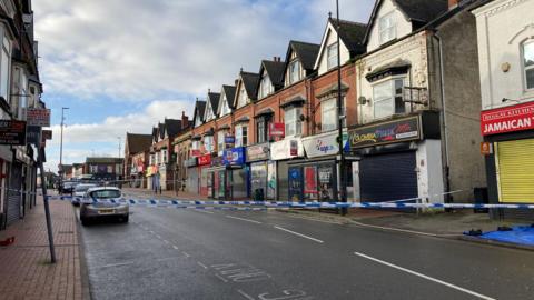 A police cordon stretches across a high street. Dozens of shops and a few cars can be seen parked on the side of the road.