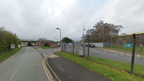 An image taken from the road that passes the military base. There are wired fences running alongside the base with two large gates opened at the main entrance. The image has been taken on a cloudy day.